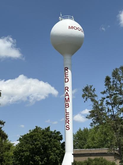 A white water tower with a spherical top displays the words "MOOSIC" and "RED RAMBLERS" in bold red letters. It stands against a blue sky with fluffy clouds, surrounded by trees.
