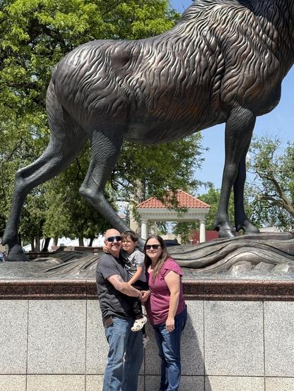 A family stands in front of a large horse statue in a park. The group includes a man, a woman, and a child, all smiling. The scene is captured on a sunny day with greenery in the background.