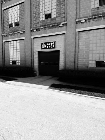 A black and white image of an auto shop entrance, featuring a sign that says "AUTO SHOP" above a dark garage door. The structure has large windows and is surrounded by neatly trimmed bushes and a concrete walkway. A portion of the road is visible