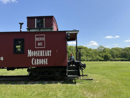 A person sits on the steps of a red train caboose labeled "Mooseheart Caboose" against a backdrop of green grass and trees under a clear blue sky.