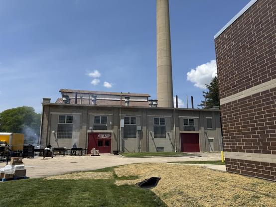 An industrial power plant building with a tall smokestack, featuring red doors and multiple windows. In the foreground, there are tables and equipment, suggesting an outdoor event or setup. The sky is blue with some clouds.