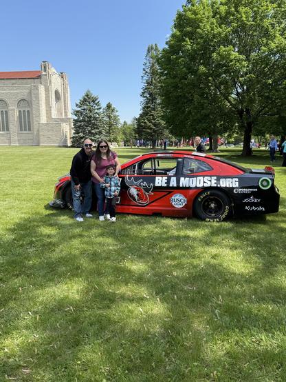 A family of three, including a woman, a man, and a young boy, poses together beside a race car parked on grassy grounds. The car features a red design with "BE A MOOSE.ORG" prominently displayed on the side. 
