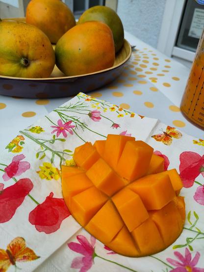 Zu sehen ist eine halbierte und in kleine Würfel geschnittene Mango, die auf einer Serviette liegt. Die Würfel sind noch mit der Fruchtschale verbunden. Die Serviette hat verschiedene Blumen und Schmetterlinge als Motiv ausgedruckt. 
Im Hintergrund sind mehrere Mangos in einer runden Schale zu erkennen. 
