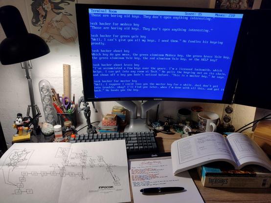 Infocom's 'Lurking Horror' on a computer screen, notes, maps and manual visible on the desk.