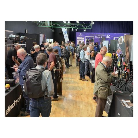 A busy exhibition hall filled with attendees exploring various photography booths and displays, with several people closely inspecting gear and speaking with vendors