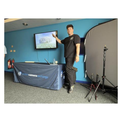 A presenter standing beside a table with a Coastal Camera Exchange banner, gesturing toward a screen during a live demonstration or talk in a seminar room