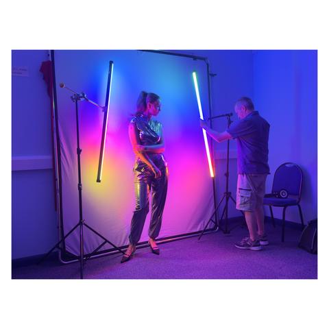 A colorful lighting setup in a darkened room, with a model posing between vertical neon lights and an assistant adjusting the scene, casting vivid hues across the backdrop