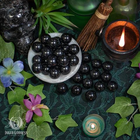 Several specimens of the "Obsidian Mini Sphere" product are arranged on a dark lace-covered surface. Several more specimens fill a selenite bowl. They are less than 1 inch across and highly polished. Presented by Inked Goddess Creations.
