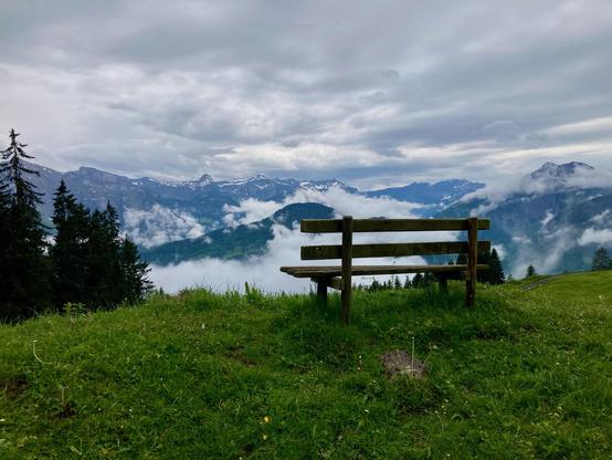 Bank in einer Wiese. Blick Richtung Alpenkette, die Gipfel teilweise mit Schnee. Viele Wolken im Bild, regnerisches Wetter.