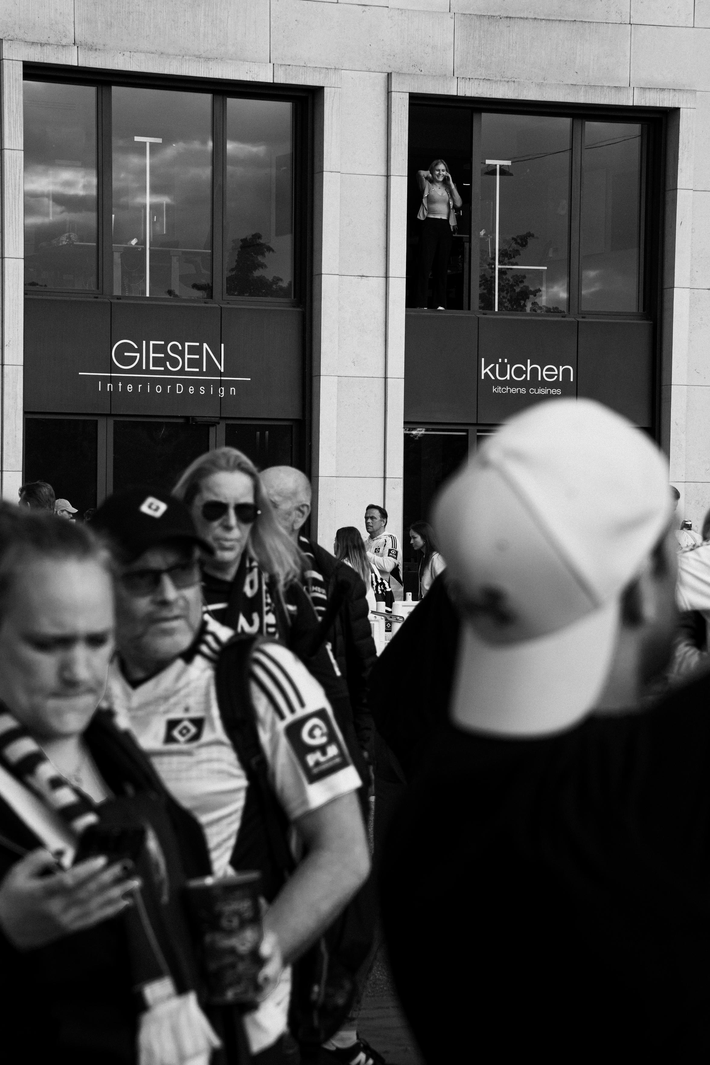 Photo showing a group of football fans in the foreground and a woman standing in a large window in the background.