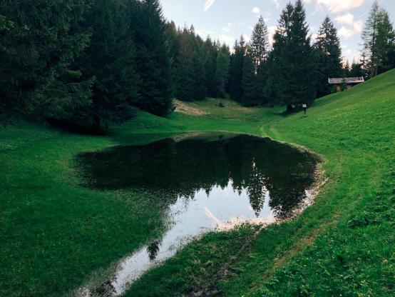Un prato in montagna con una casetta sullo sfondo ed un piccolo laghetto al centro