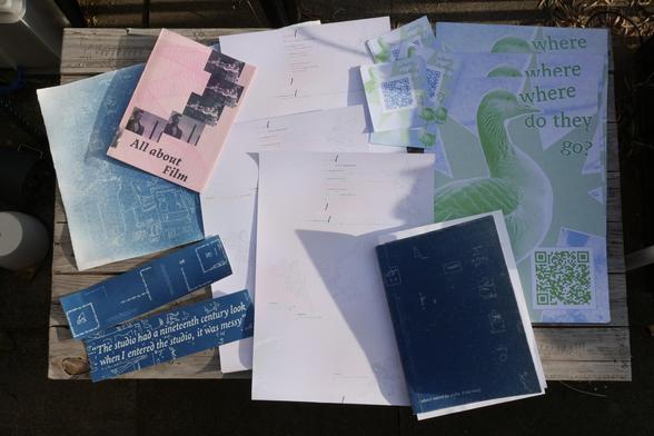 Table with a stack of several items produced today, including a cyanotype print, a booklet with a transparent sheet over a pink cover, bound using stitching, three posters with a combination of generative art and the code that generated it, some posters with a goose and the words "where do they go?" and a QR code, some small QR code sheets, two bookmarks made using cyanotype and a perfect bound book with Cyanotyped cover