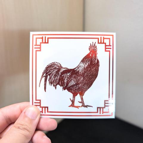Photo of a left hand holding a square sticker. The sticker is a holographic drawing of the Chinese Zodiac animal: the Rooster.