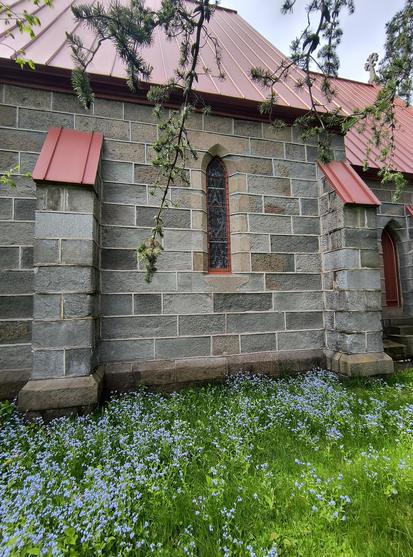 Costado de una vieja iglesia de piedra con techo rojo, puerta y una ventana roja al centro. Se encuentra sobre un terreno con pasto lleno de florecitas azules o tal vez moradas.