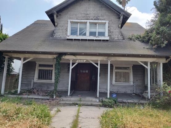 A slightly tilted picture of a decrepit abandoned house owned by musician Glenn Danzig. The house is dirty with a small pile of bricks on the porch. The lawn is overgrown and everything is dingy.