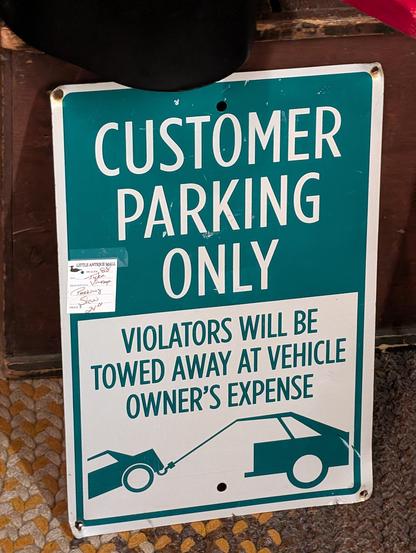 "Customer Parking Only" sign. The picture is intended to show a car being pilled by a tow truck, but only the back of the truck is visible, making it look like the backwards facing half of a split larger car with a tow cable attached to the end of the roof.
