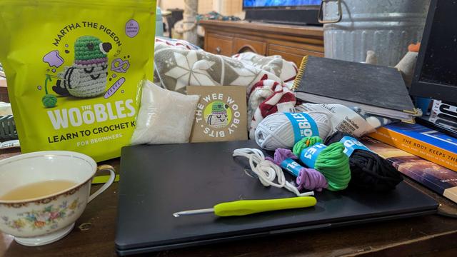 A picture of a messy desk: white, purple, green, black,, and gray yarn are bundled together with a green crochet hook. It's a Martha The Pigeon woobles kit, prepared to be made.