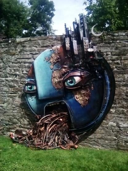 Sur le mur en pierres d’un parc, une sculpture ronde, la terre (on devine l’Afrique, l’Europe et le bleu de l’Atlantique) est affublée d’un visage : deux yeux et une bouche grande ouverte qui vomit des cables. Des gratte-ciels poussent au dessus de l’œil droit.