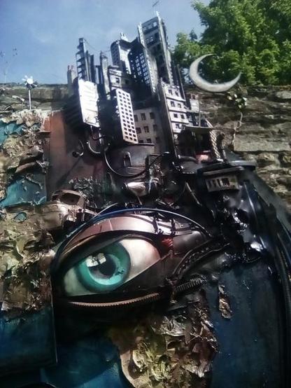 Photo en gros plan de l’œil droit de la sculpture et des gratte-ciels. On voit qu’ils sont fait de matériaux de récupération, grilles, plaque d’immatriculation… Ils sont surmontés d’un croissant de Lune.