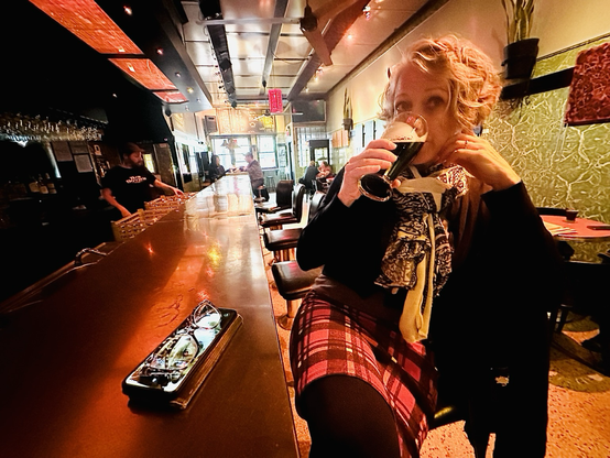 A woman sitting at a red-lit bar drinking a pint.