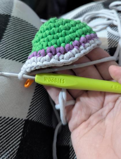 The first nine rounds of the crochet kit "Martha the Pigeon" by the Woobles company. It's a green dome of a street pigeon's head with a purple layer at the base of the neck transitioning into gray. A hand is holding the crochet project with a chartreuse crochet hook still attached.