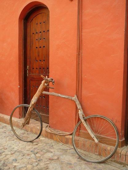 Bicycles leaning against the wall of a building with ochre plaster, on a cobbled road, nest to a rustic but well kept door. The bicycle has spoked wheels bu the frame is made of what appears to be sticks, as if they were grown in the shape of a bicycle frame. Artist unknown, location unknown.