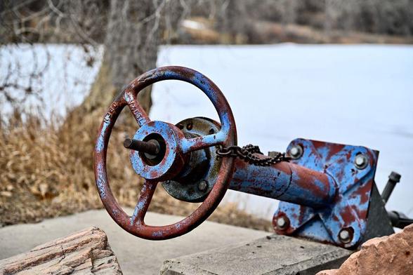 Close-up of a rusted red and blue industrial valve mounted to concrete by a body of water. The wheel is weathered, chained in place, with dry grass and a tree trunk in the background.