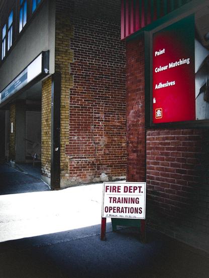 A white sign sits near the entrance of an alley reading "Fire Dept. Training Operations". The bricks  of nearby buildings are seen as well as sunlight streaming from the alley.