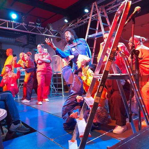 Die Schauspieler und Schauspielerinnen des Stuttgarter Interkulturellen Ensembles (SIE) posieren mit unterschiedlichen Gesten und Gesichtsausdrücken auf der Bühne zwischen mehreren Leitern. Die Leute tragen Arbeitsoveralls in den Farben Blau, Gelb und Pink. An den Seiten der Leitern befindet sich ein schmaler Klebestreifen mit weißen LEDs. Die Bühne wird mit blauem Licht beleuchtet. Deshalb ist auch der Boden blau. Am rechten Bildrand steht hinter der Leiter ein Mikrofon. Am linken Bildrand ragen die Beine eines Zuschauers ins Bild.