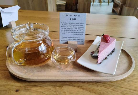 A moment in tearoom. A tray on a table with a glass pot of white tea, a small glass cup, and a slice of strawberry-white chocolate cake.