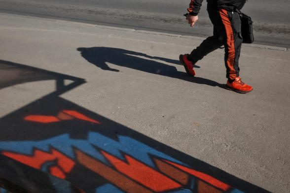 On the asphalt falls the shadow of the stop and red and blue reflexes from the stained glass panels with which the stop is decorated. A man in red sneakers walks by. Only his feet are visible. A shadow falls from the man.