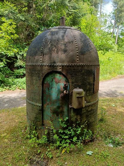 A small cylindrical rusty metal hut with a domed roof. It is made out of separate panels riveted together. There is a chimney poking out the top and a metal door in the front with a curved top. The door has a metal handle and some kind of lock. There is a small metal tank bolted to the front. The hut is surrounded by trees and grass.