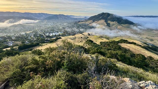 A descending grassy hill is replaced by urbanization on one side and more grassland on the other. Mountains and fog linger. 