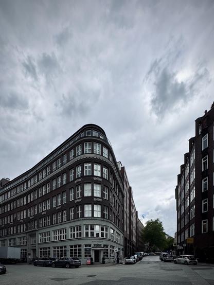 Eine Straßenszene mit einem markanten, gebogenen Eckgebäude mit zahlreichen Fenstern. Das Gebäude ist mehrstöckig und aus roten Ziegeln gebaut. Autos sind entlang der Straße geparkt und der Himmel darüber ist bewölkt.