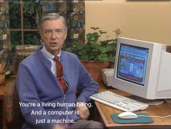 Screenshot of the Mister Roger's Neighborhood show. Mister Rogers sits in front of a mid-1990s-style PC, looking at the camera as he says, “You’re a living human being. And a computer is just a machine.”