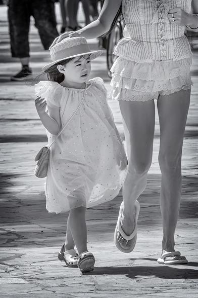 Mother and daughter walk along Waikiki Beach in Honolulu, Hawaii. Daughter is wearing a sun hat with a ribbon and and wide brim and mom is holding it on her head to stop it from being blown away in the wind.