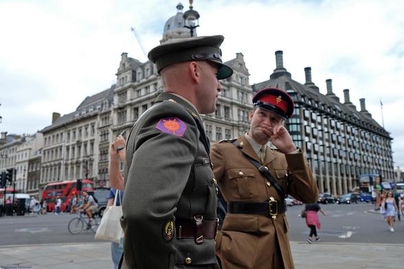 Photograph by Fenichel:
2 soldiers, wearing different uniforms (olive on left, with bright purple-red soldier patch, and brown uniform with shoulder strap, red hat and wide belt on right).

They are standing in a square in London, with a red double-deck bus, bicyclist, and pedestrians in the background. The one on the left listens intently as the one on the right says something with a finger in front of his lips. They look cordial.
