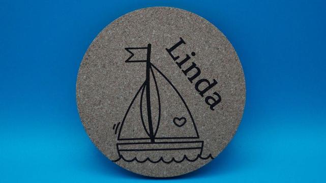 Runder Korkuntersetzer mit graviertem Motiv eines Segelbootes, daneben der Name „Linda“, schräg eingraviert.