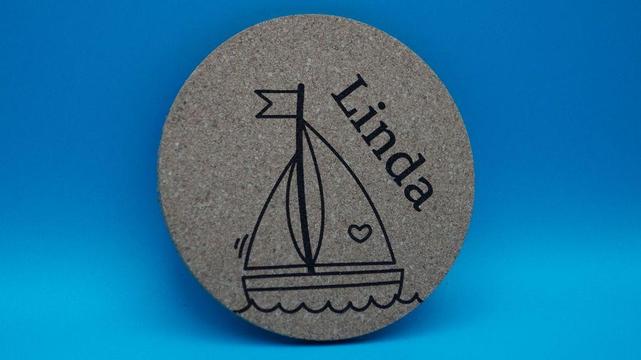 Korkuntersetzer mit Segelboot- und Herzmotiv, personalisiert mit dem Namen „Linda“, vor blauem Hintergrund.

