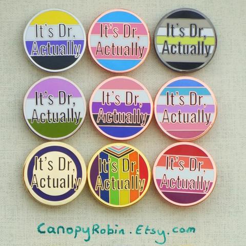 Enamel pins that say "It's dr, actually" on trans, nonbinary,  agender, genderqueer, genderfluid, bigender, intersex, progress, and lesbian pride flags. There are also versions on black, white, hot pink, purple, dark green, light pink, light blue, and light green colour backgrounds