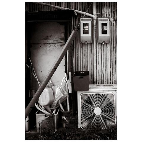 In this black and white photograph, an old corrugated tin wall hosts a tableau of urban disorder. A plastic bin overflows with watering cans and umbrellas—two of which are grouped together, while one stands separately, propped against a door. A weathered air conditioning unit sits below, its fan grill adding a geometric contrast.