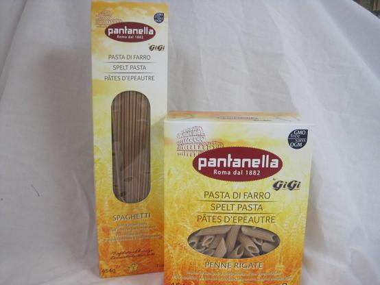 The image shows two boxes of pasta from the brand "Pantanella," with the tagline "Roma dal 1882." The box on the left contains "Spaghetti" made from spelt, labeled in Italian as "Pasta di Farro," English as "Spelt Pasta," and French as "Pâtes d'Epeautre." The box on the right contains "Penne Rigate," also labeled in the same languages. Both boxes are predominantly yellow with a sunburst design and feature a window showing the pasta inside. The "Spaghetti" box is taller and has a weight of 454 grams, while the "Penne Rigate" box is wider. Both boxes have a "GMO free" label, indicating that the product does not contain genetically modified organisms. The background is a plain white fabric, providing a neutral backdrop for the products.
