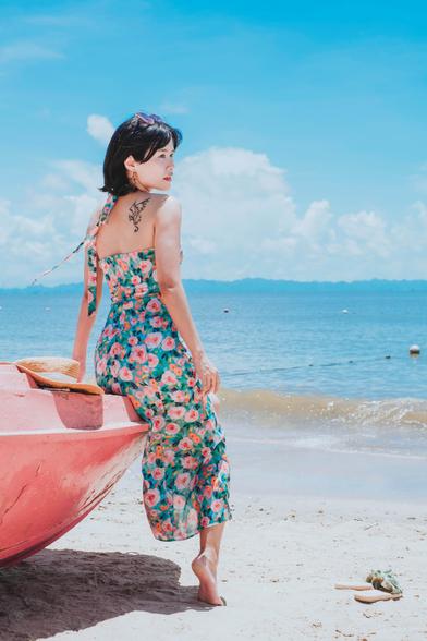 Asian woman in a flower dress with a dragon tattoo on her back leaning against a red boat on a beachfront