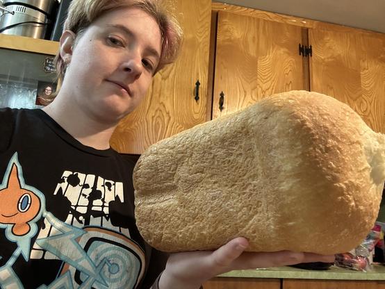 Short haired white person in a black t shirt featuring the Pokemon Rotom holding up a fresh baked loaf of white bread. 