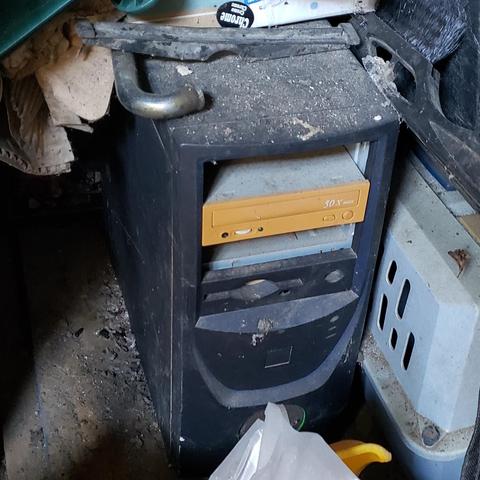 Photo of an old computer freshly uncovered beneath a pile of stuff in a garage. The system was painted black with neon green accents. A wire mesh covered hole can be seen on the front toward the bottom of the case peeking out from behind a plastic bag. A very yellowed CD-ROM drive and the door to a floppy drive are visible in the mostly stripped machine.