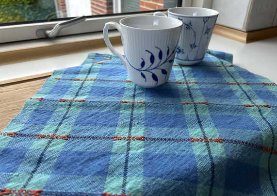 A blue checked handwoven teatowel sitting on a wooden cutting board. Two blue and white mugs sit on the teatowel