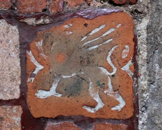 A very worn clay tile with a white-ish outline of a griffin with one of its front paws, its wings and its tail all raised. Much of the design, especially the head and the front of the body are almost worn away. Either side of the griffin are brackets for decoration.