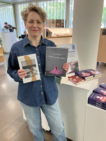 Farkkuasuinen kirjailija seisoo kirjaston aulassa käsissään kaksi kirjaa, tekijät Annie Ernaux ja Liv Strömquist. Vieressä pöydällä Margit-romaaneja.