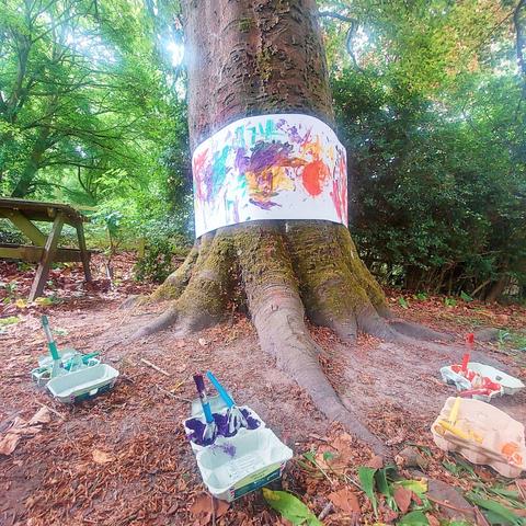 One of the older trees in the museum gardens, with a roll of paper wrapped around its thick trunk. The paper has been daubed with colourful paint, and you can see the egg boxes with paint and brushes at the tree's base. The entire area is well-shaded by the tree canopy.