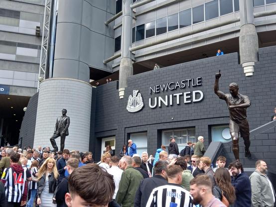 Crowd outside St James' Park with the statues of Sir Bobby Robson and Alan Shearer in view.